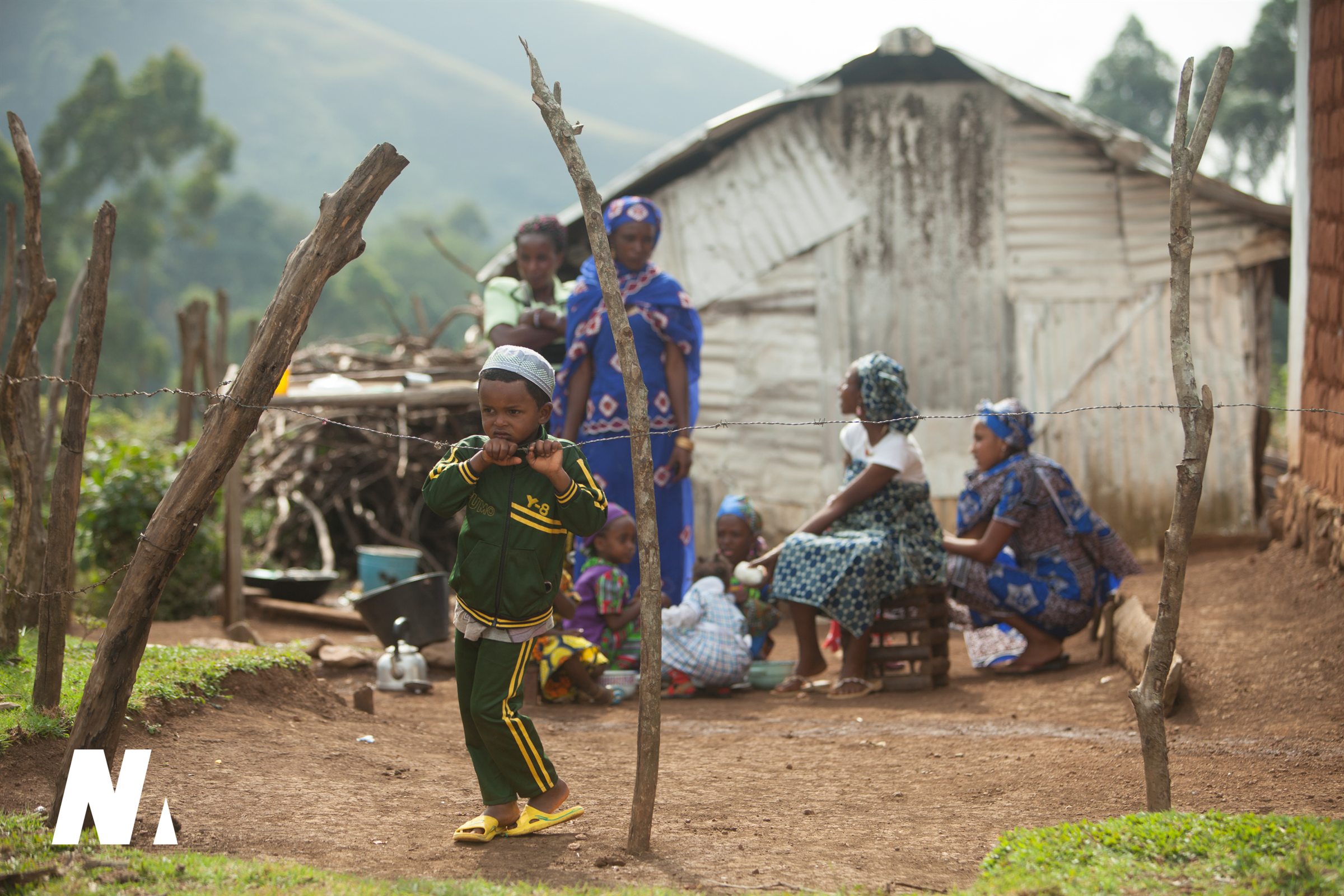 A child stands at a simple fence with family gathered behind